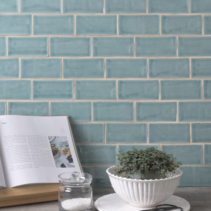 Wall of a pale green blue medium brick metro tiles with beige grout against a stone worktop with a cookbook and herb planter