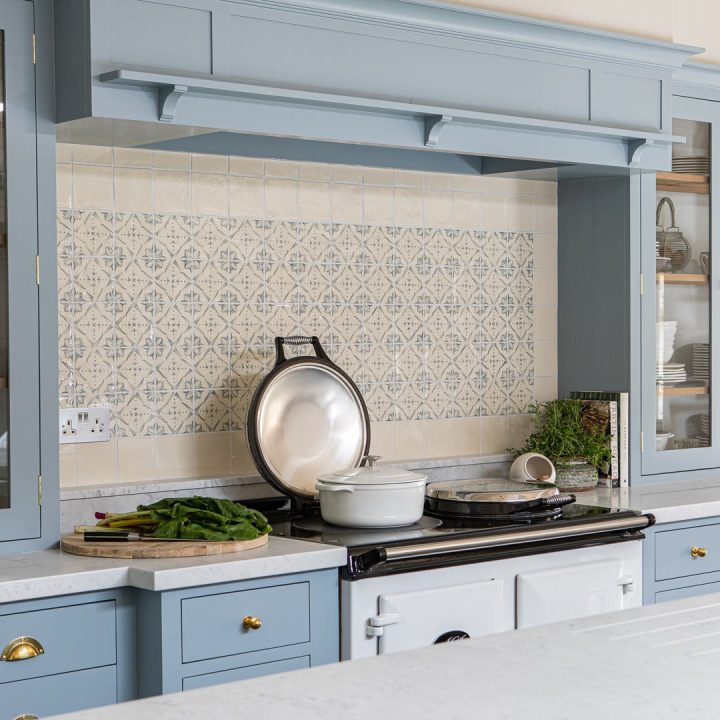 Wall of powder blue Mediterranean pattern hand painted tiles with a border of plain tiles behind a white aga and blue kitchen cabinets