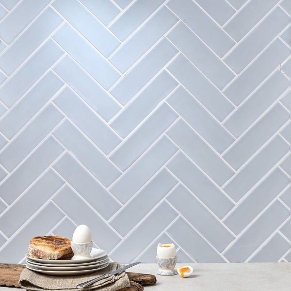 Wall of skinny pale blue matt metro tiles with silver grey grout laid in a herringbone tile pattern styled with a pile of plates
