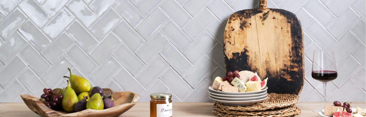 Small brick light grey Savernake College Fields wall tiles in a herringbone pattern on a kitchen wall behind a fruit bowl, wooden platter and glass of wine