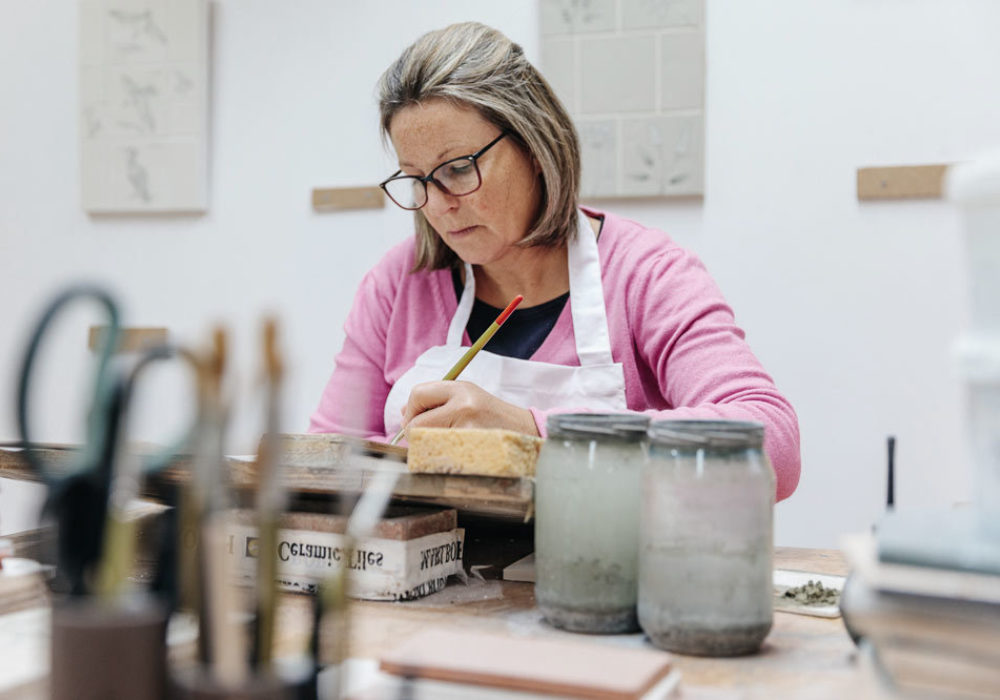 Kirsty hand painting a tile at her desk with tile panels in the background and her paints in the foreground.