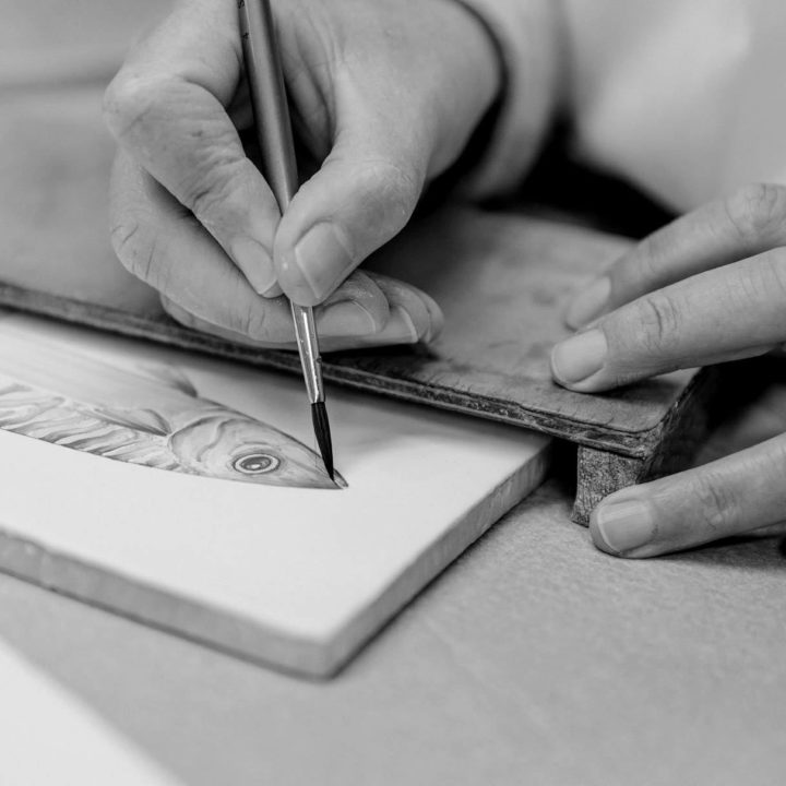 Black and white image of a close up hand of a hand painter painting a fish tile