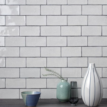 Wall of aged crackle glazed brick tiles with grey grout behind a vase white vase and green bowl