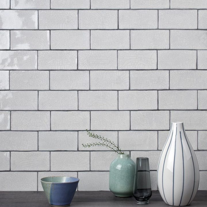 Wall of aged crackle glazed brick tiles with grey grout behind a vase white vase and green bowl