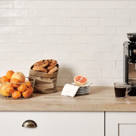 Wall of skinny warm white tiles with white grout against an oak worktop behind a coffee pot and fruit bowl