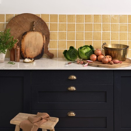 Wall of handmade saffron yellow square wall tiles behind a white worktop and navy kitchen cupboards