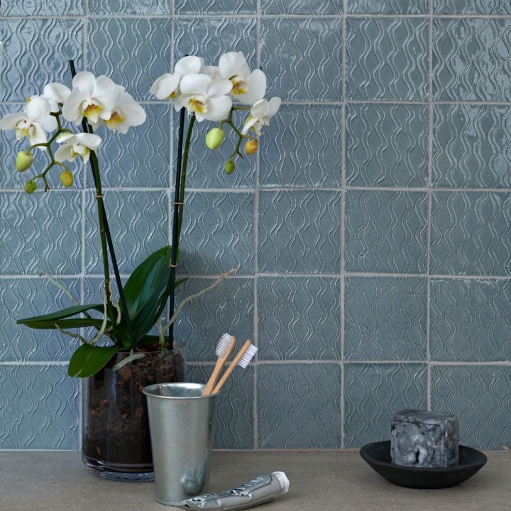 Wall of artisan rustic handmade subtle blue square tiles with a hand piped pattern with beige grout behind a vase of orchids