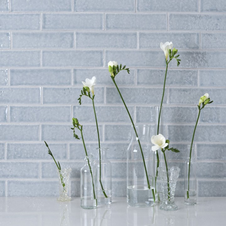 Wall of skinny blue tiles with silver grey grout behind vases of white orchids on a grey worktop