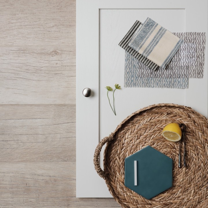 Flatlay of oak floor planks with a white kitchen cupboard door, a blue hexagon tile on a rattan tray and fabric swatches