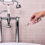 Traditional chrome bath taps in front of blush pink matt metro tiles with a hand pouring bubble bath.