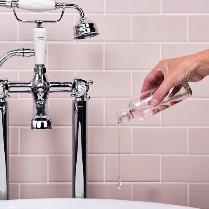 Traditional chrome bath taps in front of blush pink matt metro tiles with a hand pouring bubble bath.