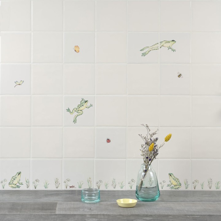 Wall of ivory tiles with hand painted sitting and leaping frogs, pond grasses and insects styled with a grass and glasses
