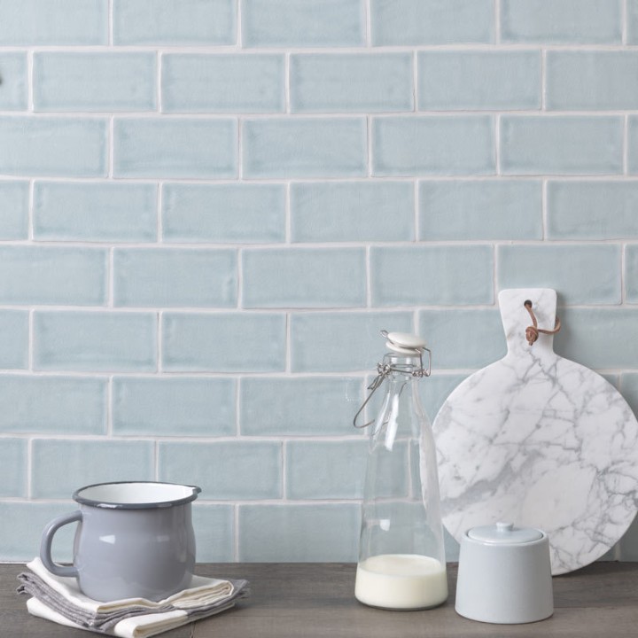Wall of pale blue medium brick metro tiles with silver grey grout against a wood worktop with kitchen accessories on top