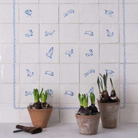 Panel of delft tiles with animal illustrations framed with a blue border. Three pots of spring bulbs are in front